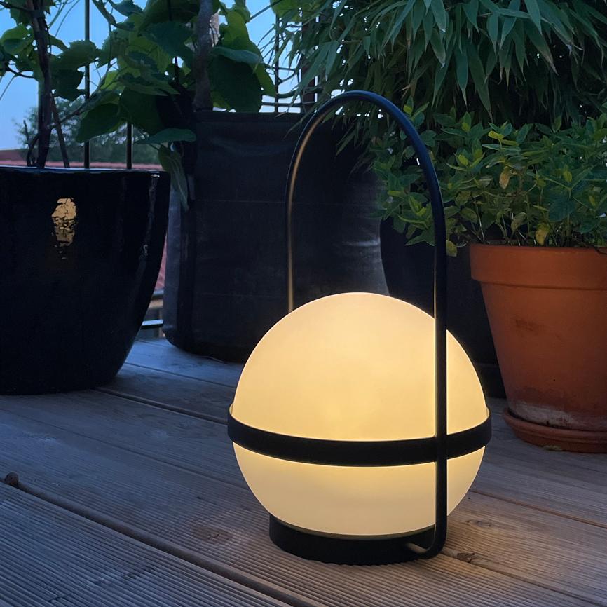 Sfærisk bærbar lampe lyser med varm glød; den står på en træterrasse mellem terracotta- og sorte plantekrukker med grønt løv i skumringslys.