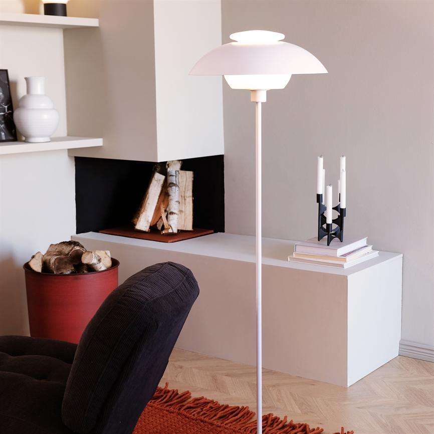 Gulvlampe står tændt ved en lav hvid bænk med bøger og lysestage; en lille pejsåbning med stablet birkebrænde og en mørk sofa samt rødt tæppe i forgrunden.