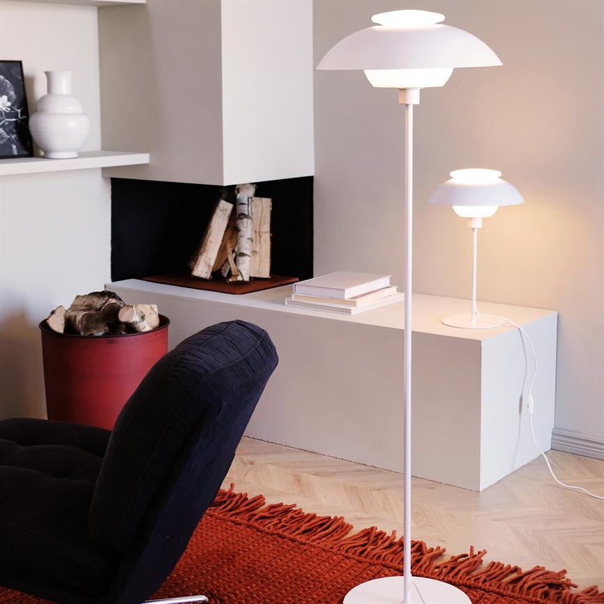 Hvid gulvlampe står og lyser ved en lav hvid reol; mindre bordlampe, stabler af bøger, indhak med birketræ, en mørk lænestol og rød tæppe i et lyst, moderne opholdsområde.