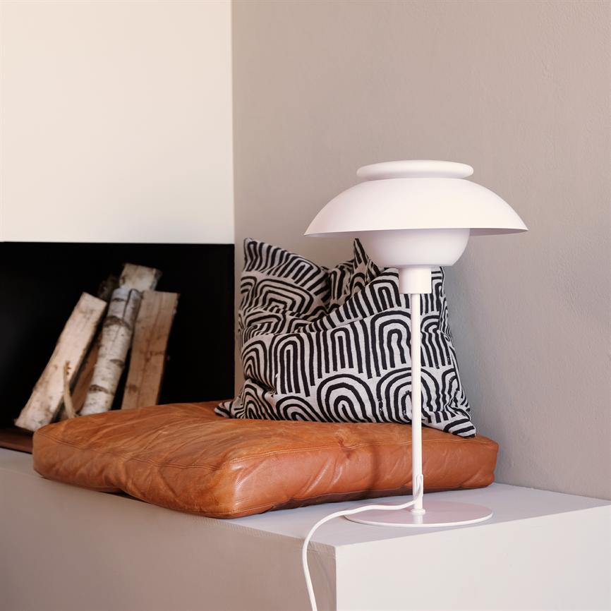 En hvid bordlampe står på en hvid hylde; ved siden af ligger en brun læderpude og en sort-hvid mønstret pude, foran en ildstedsåbning med stablede birkestammer.
