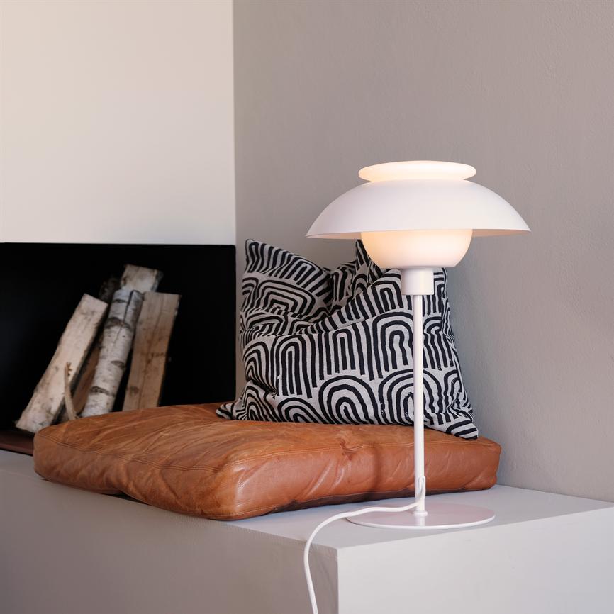 En hvid bordlampe lyser blødt på en hvid hylde ved en væg; foran en brun læderpude og en sort‑hvid mønstret pude, med birketræsstammer i en nærliggende niche.