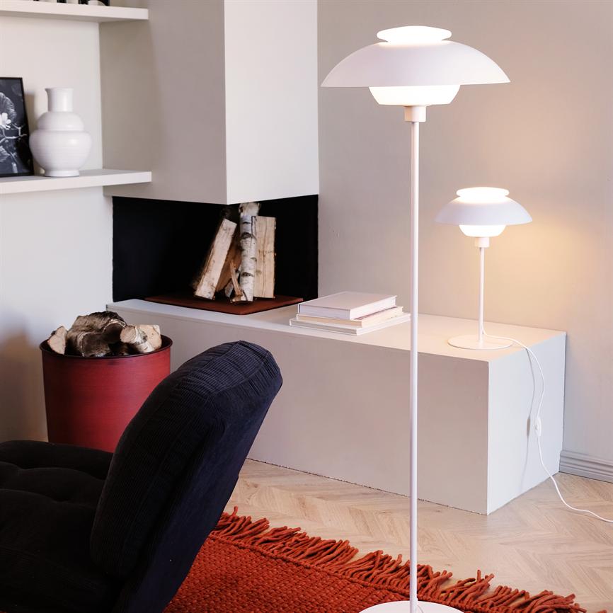 En hvid gulvlampe lyser blødt; en mindre matchende bordlampe står på en lav hylde. Omkring: indbygget pejs med brænde, rød brændekurv, mørk lænestol og rødt tæppe.