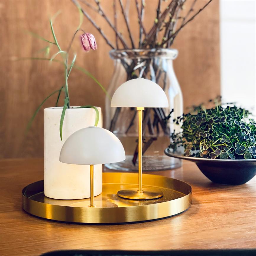 To små hvide bordlamper står på en rund guldfarvet bakke, udstråler blødt lys; omgivet af en hvid cylinder, en glasvase med kviste og en grøn plante på et træbord.