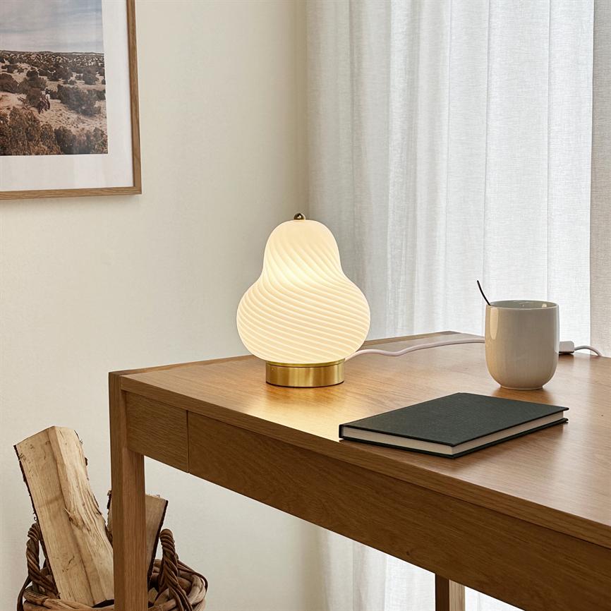Bordlampe med hvidt, bølget glas står tændt på et træbord og udsender varmt lys; ved siden af ligger en lukket notesbog og et krus foran lyse gardiner og vægkunst.