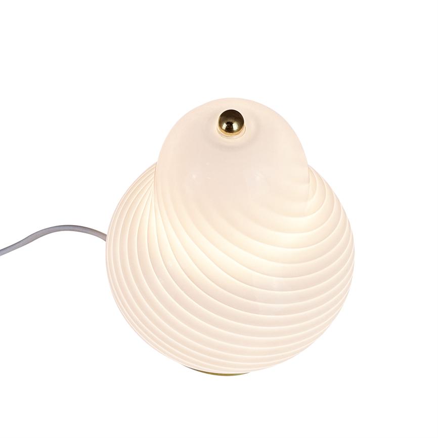 Rund bordlampe i opalglas udsender blødt, varmt lys; ribbet overflade, lille messingknap på toppen og synlig grå ledning på hvid baggrund.