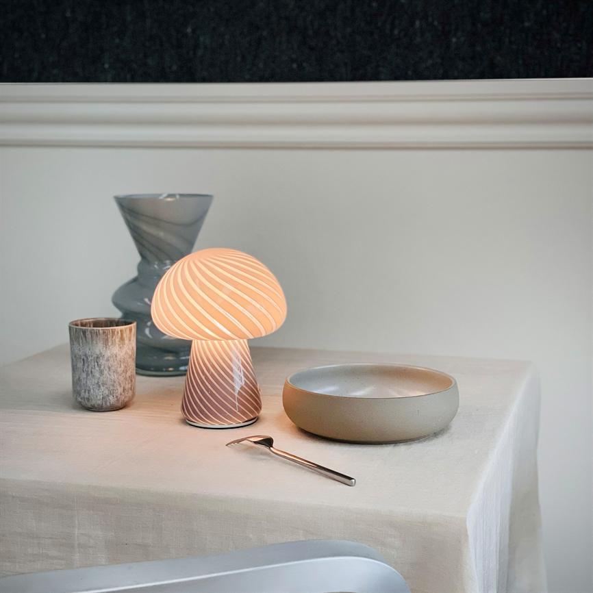 Svampeformet bordlampe udstråler varmt lys på et dækket bord; den står ved en skål, en ske, et krus og en vase på en lys dug foran en lysegrå væg.