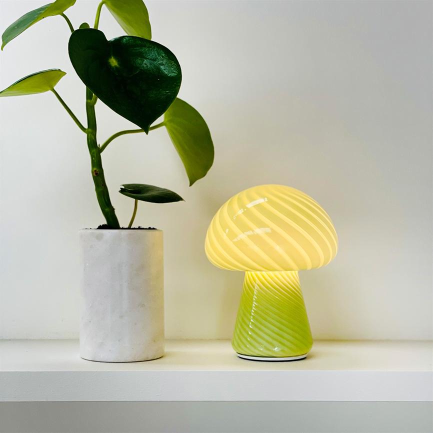 Svampeformet lampe udsender varmt, gult lys; den står på en hvid hylde ved siden af en grøn potteplante i cylindrisk hvid potte foran en ensfarvet hvid væg.