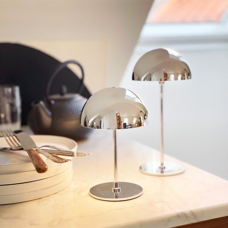 To krombelagte bordlamper står og spejler rummet på et marmorbord; omkring dem ligger stablede tallerkener, bestik og en sort tekande i et lyst køkken med skråvindue.