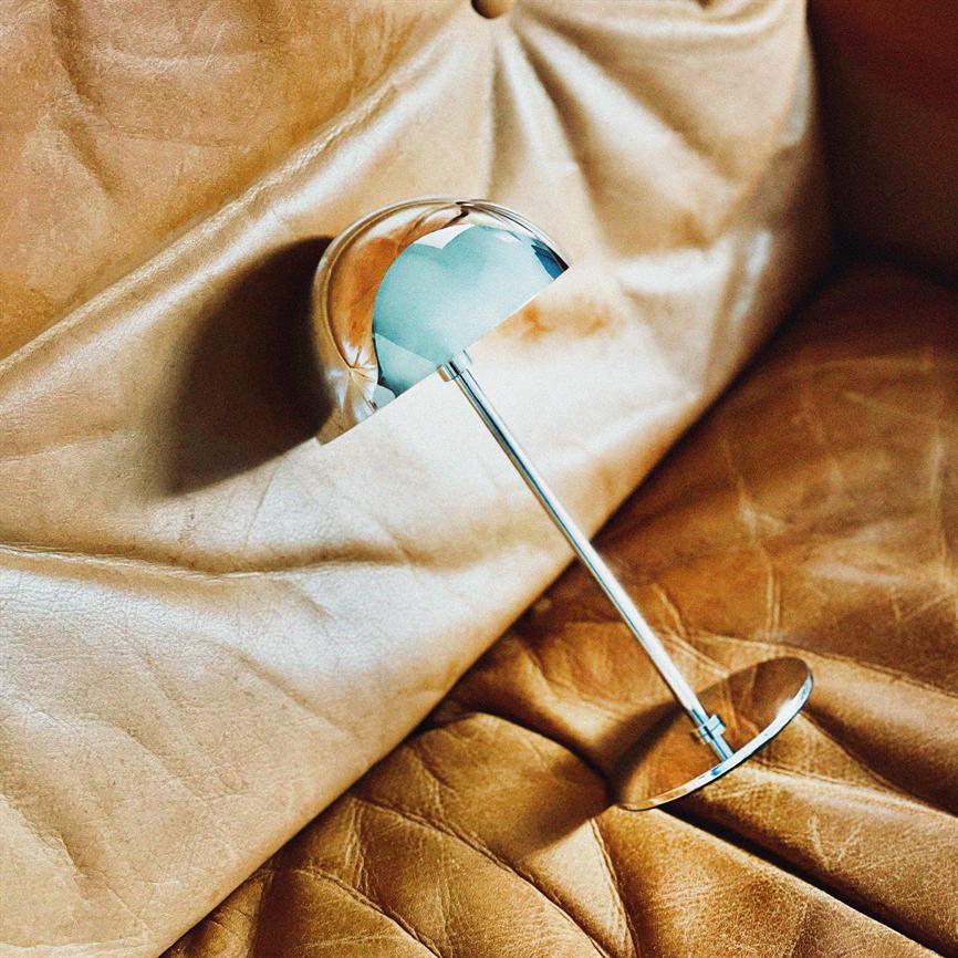 En krom bordlampe ligger på siden, let tiltet og reflekterer omgivelserne; dens slanke stålskaft og halvkugleformede skærm hviler på rynket, gyldenbrunt læder på en sofa.
