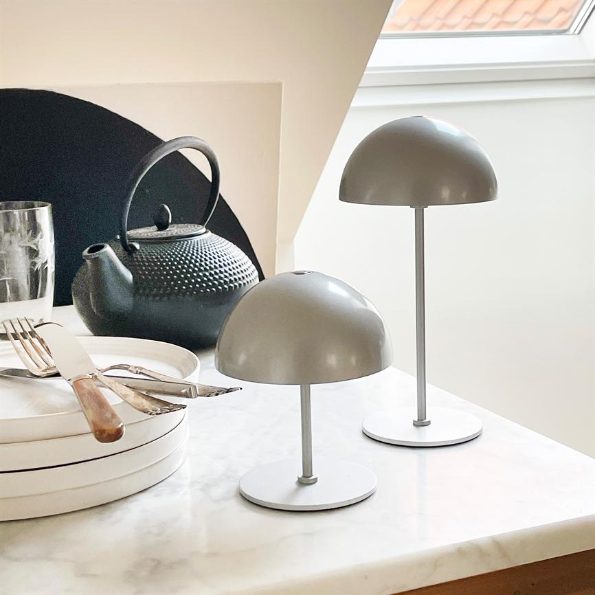 To grå, svampeformede bordlamper står på et marmorbord; i baggrunden en sort støbejerns tekande, en stak hvide tallerkener med bestik og et glas ved et skråvindue.