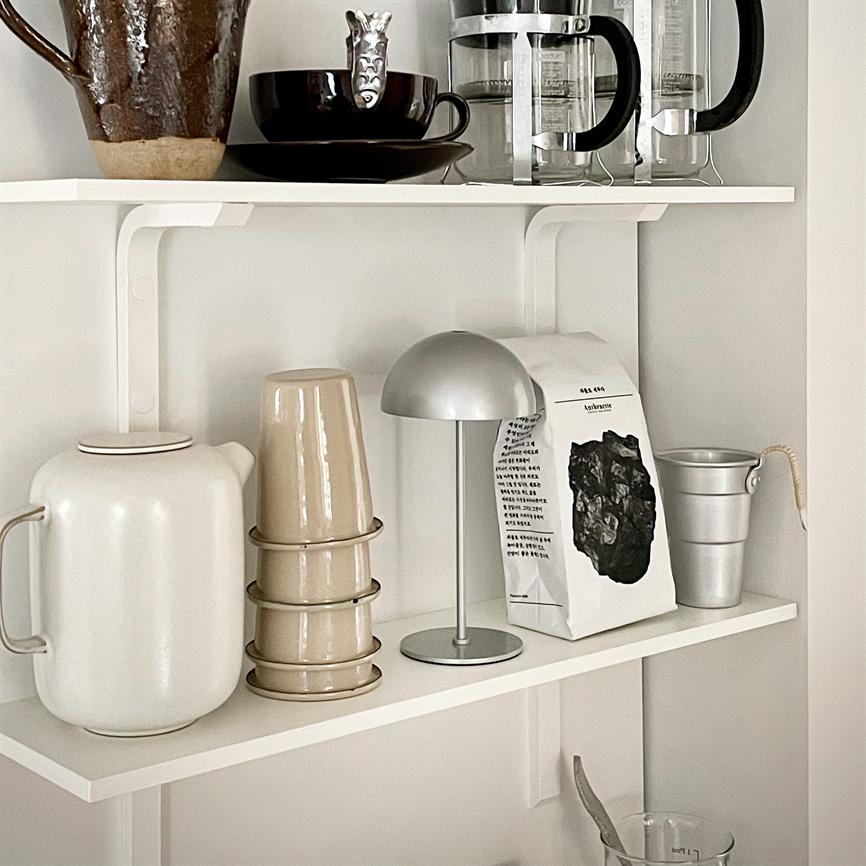 Kaffepose står midt på en hvid hylde mellem en lille sølvlampe og en metalspand; beige stabelkopper, en hvid kande og franske presskander ses på hylder omkring den.
 
 Tekst på posen: flere lodrette linjer med koreansk skrift samt et lille latinsk ord øverst — teksten er for lille til entydig aflæsning.