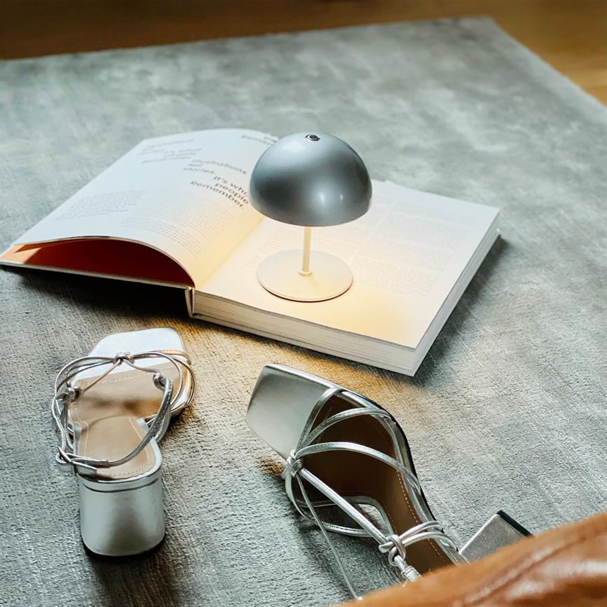 Lille grå lampe lyser på en åben bog; et par sølvfarvede, snørede højhælede sandaler ligger spredt på et lyst, blødt tæppe. Bogens tekst er ikke læsbar.