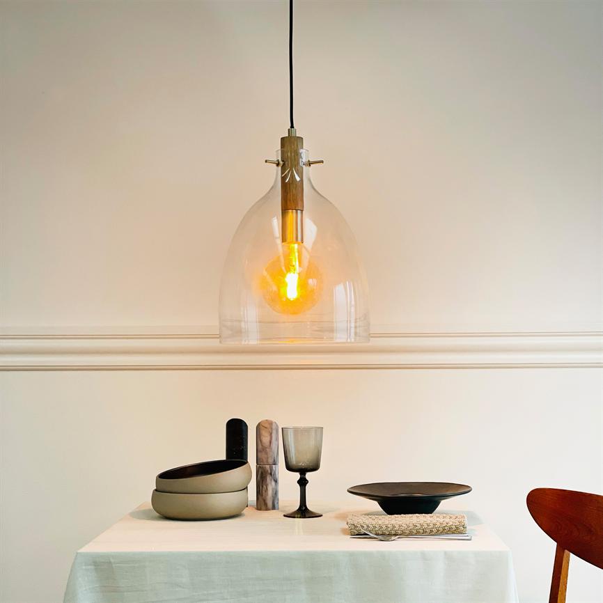 Glaspendel hænger og udsender en varm orange glød over et enkelt dækket bord med skåle, et vinglas, en sort tallerken og serviet i et lyst, minimalistisk rum.
