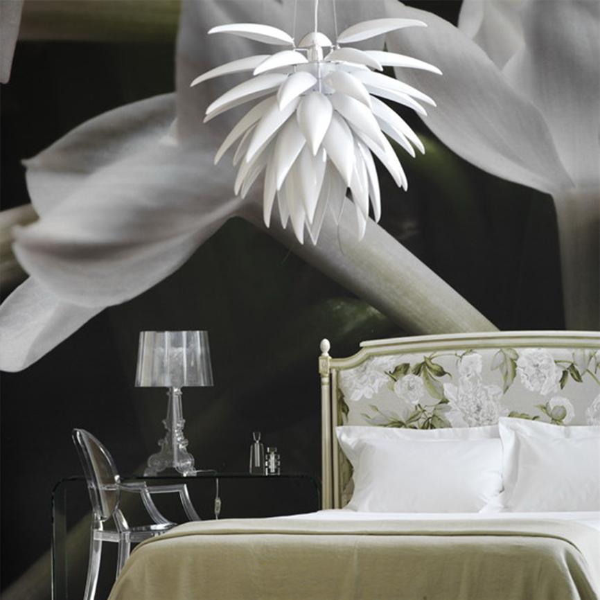 Hængende hvid blomstformet lampe svæver over en seng, sengegavl med blomstermønster, hvide puder og beige dyne, klar akrylstol og glasbord mod en mørk, stor blomsterbaggrund.