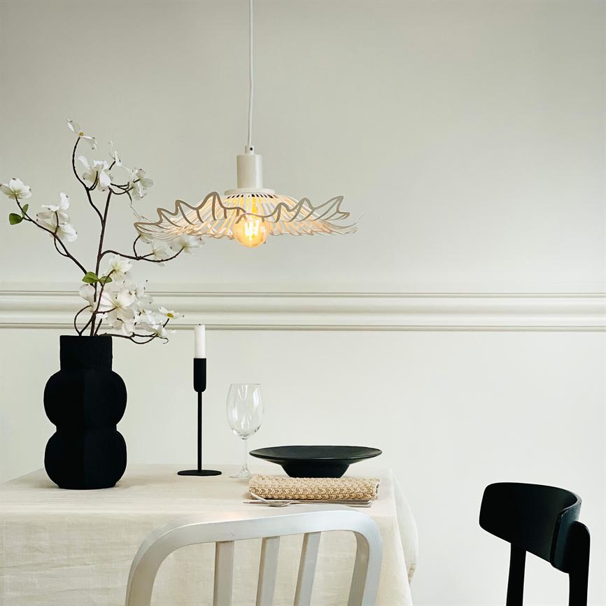 En hvid dekorativ hængelampe lyser varmt ned over et dækket bord; en sort vase med hvide blomster, ét stearinlys, vinglas og sort tallerken i en minimalistisk, lysegrå spisestue.