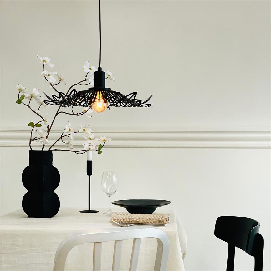 En sort flettet pendel lyser svagt over et dækket bord; en sort vase holder hvide blomster, et vinglas og en sort tallerken ligger på en hvid dug i et lyst, minimalistisk rum.