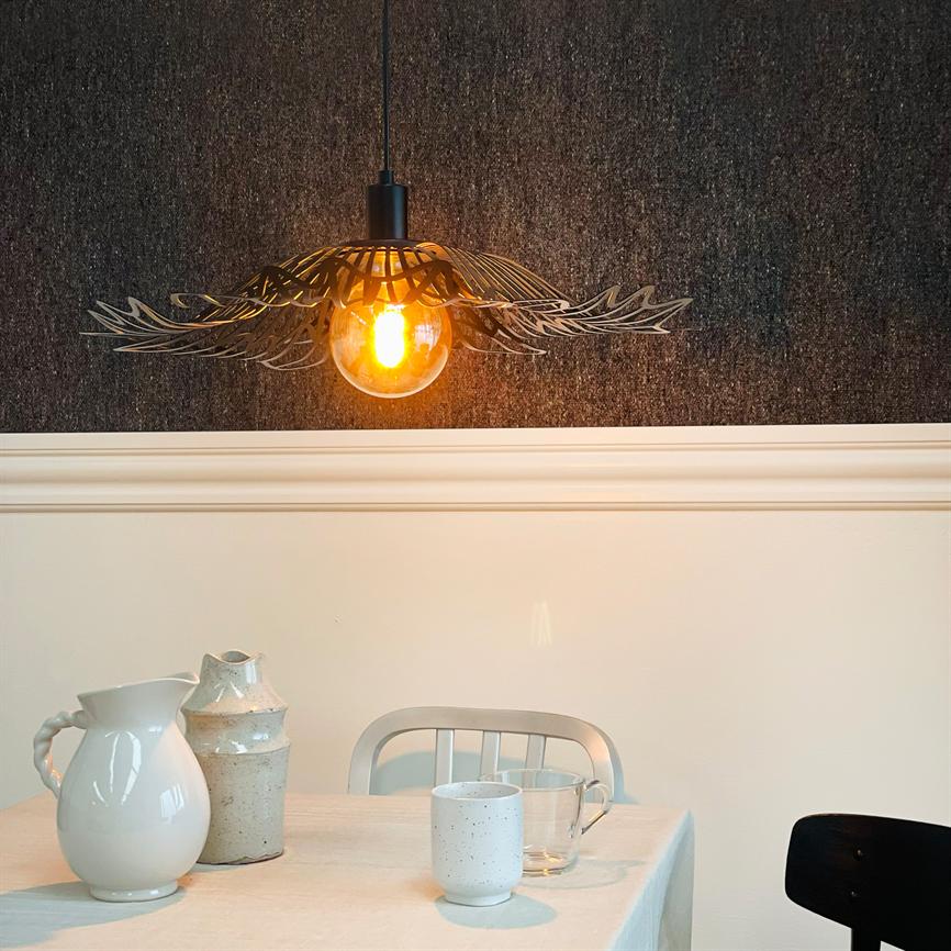 En dekorativ hængende lampe lyser varmt over et spisebord; bordet har hvide keramikkander og krus, foran hvid panelvæg og mørkt tekstureret tapet.