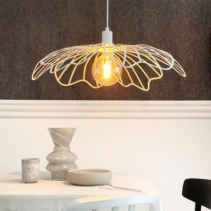 En loftlampe hænger over et bord, udsender varmt lys gennem en synlig glødepære og dekorativt metalskærm; bordet med snoet vase, krus, skål og ske foran en mørk væg.