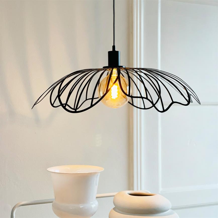 Sort, bladinspireret loftlampe hænger og kaster varm glød fra en synlig glødepære over to hvide vaser på et bord foran en lys væg og en dør.