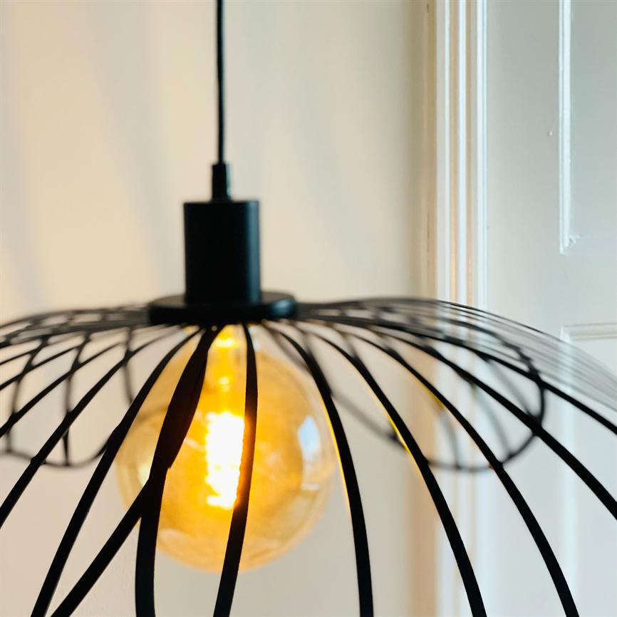 Loftlampe med synlig, glødende pære hænger i et sort, spindelagtigt metalbur og udsender varmt gult lys; omgivet af lyse vægge og en hvid dør i baggrunden.