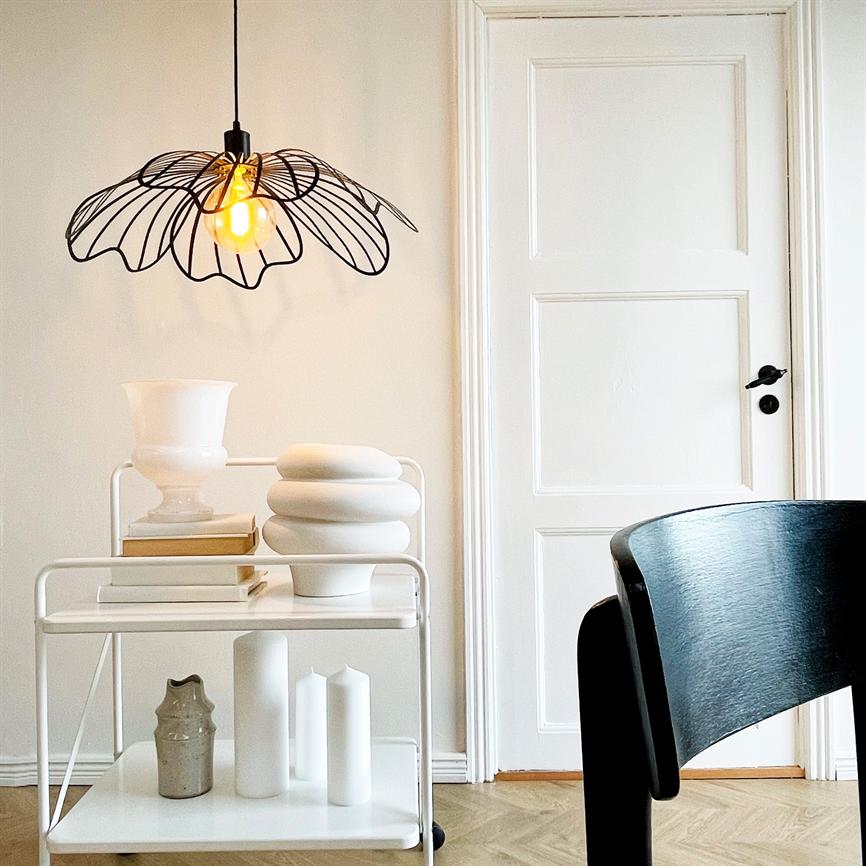 En sort, trådformet loftlampe kaster varmt, gyldent lys over en hvid vogn med keramiske vaser foran en hvid dør og en sort træstol i et lyst, minimalistisk rum.