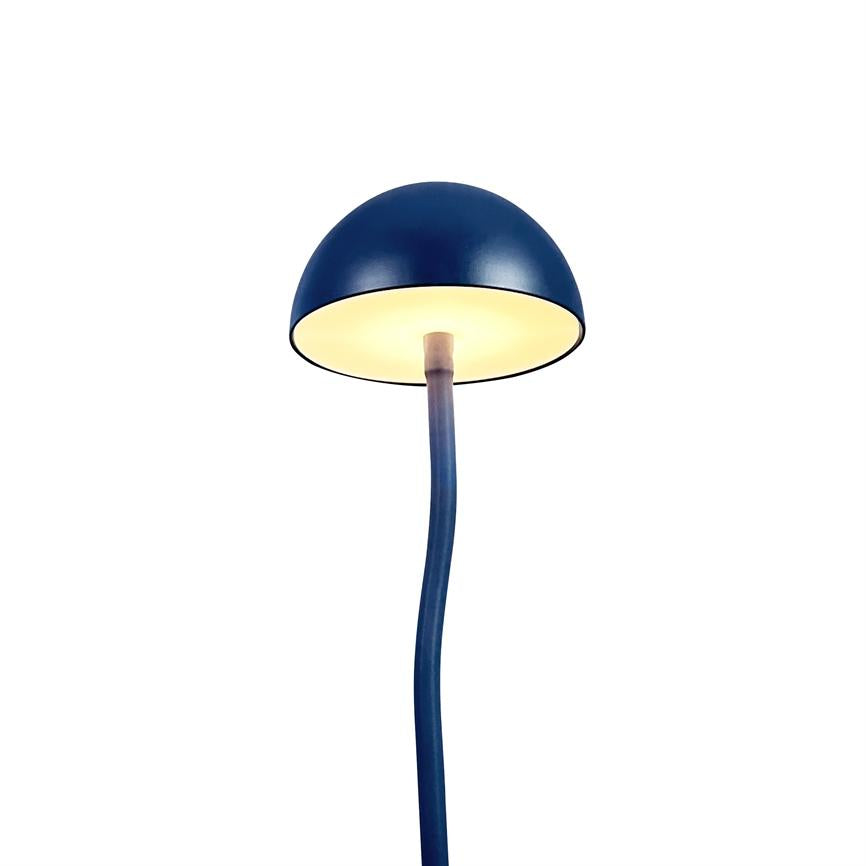 Blå bordlampe viser en halvkugleformet skærm, lyser indefra; en slank, let buet stang leder nedad mod en neutral hvid baggrund.