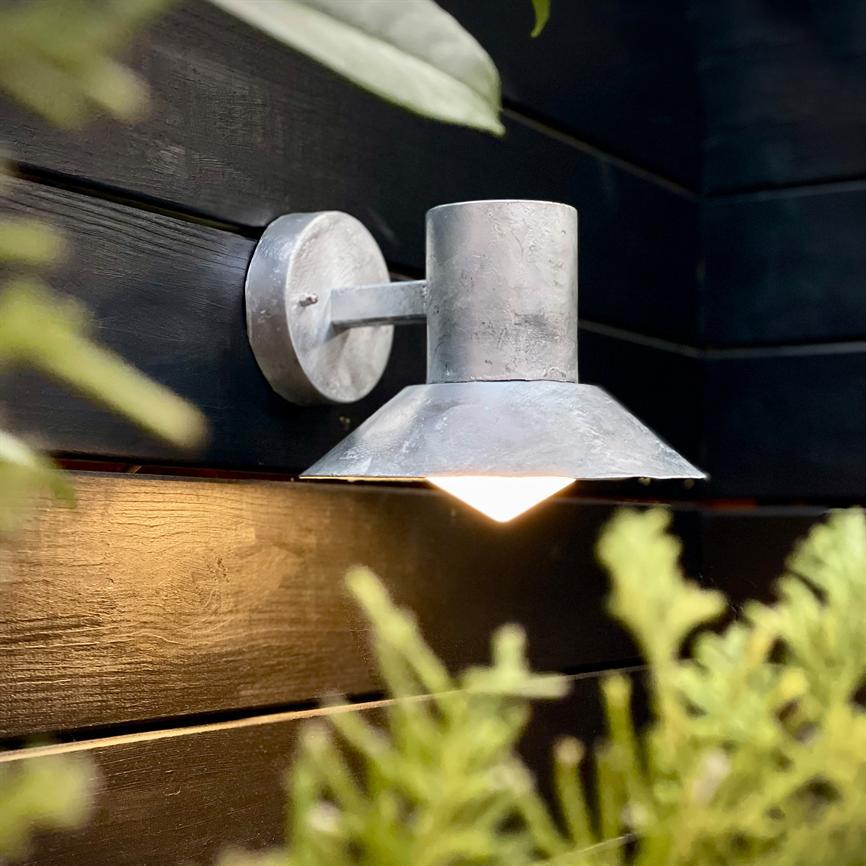 Væglampe lyser nedad; gråt metalarmatur fastmonteret på mørke, vandrette træbrædder med sløret grønt planteliv i forgrunden, varmt lys kaster glød på træets overflade.