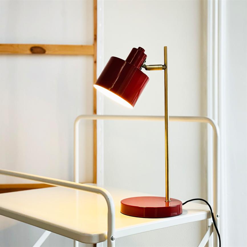 Rød skrivebordslampe lyser nedad, monteret på en messingstang; den står på en hvid metalvogns hylde med sort ledning, foran en lys væg og en enkel træramme.
