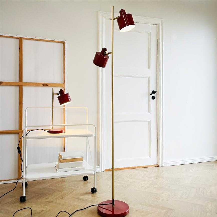 Gulvlampe i messing med to røde lampehoveder står oprejst og lyser ved en hvid dør; i forgrunden en rullende hvid vogn med matchende bordlampe og stablede bøger på lyst trægulv.
