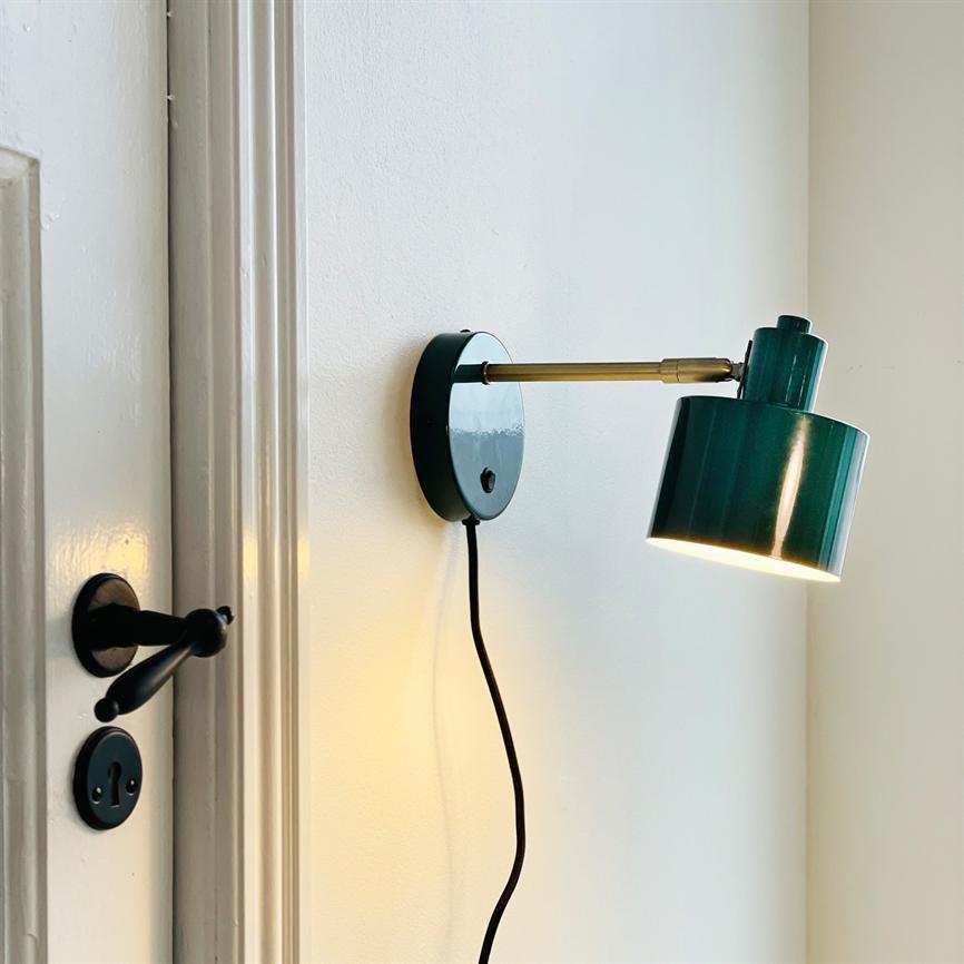 Væglampe, monteret på en hvid væg, lyser nedad; grøn metalkuppel og messingarm, sort ledning hænger ned, ved en dør med sort dørhåndtag og nøglehul.