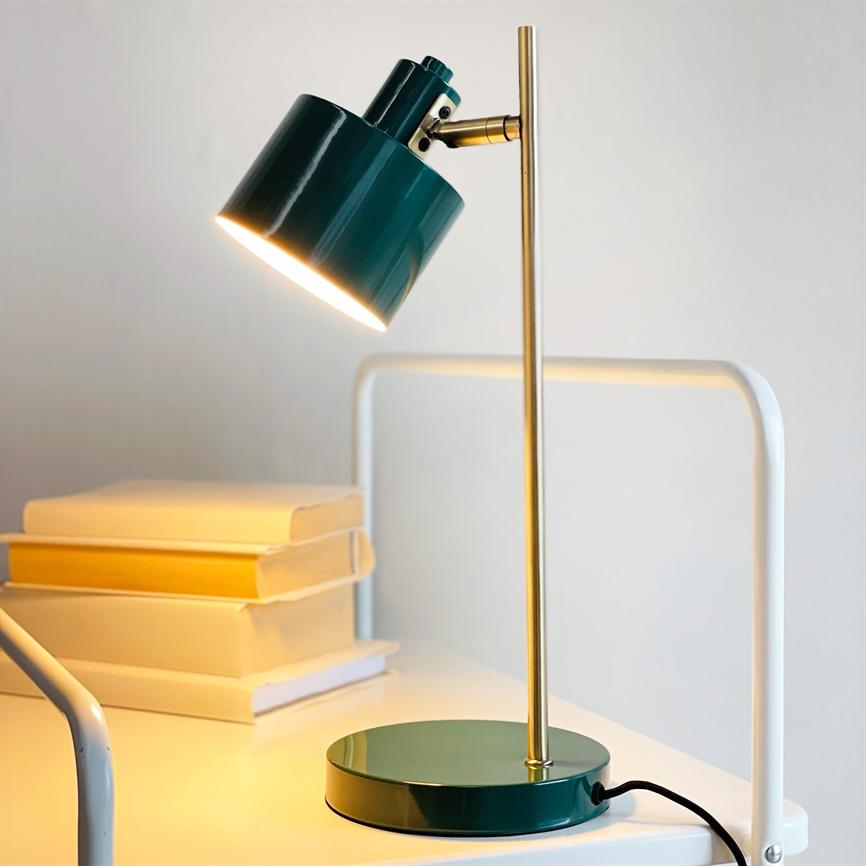 Grøn metal bordlampe med messingstang kaster varmt, fokuseret lys nedad; den står på et hvidt bord ved en stak lyse bøger foran en neutral, lys væg.