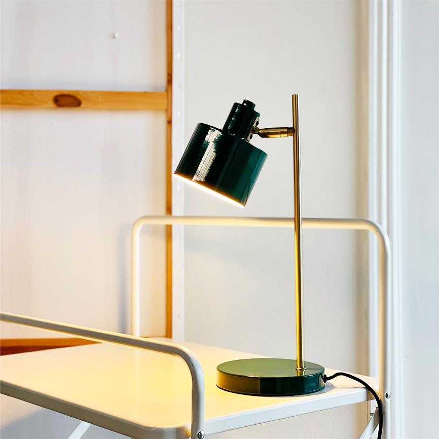 Grøn metalskrivebordslampe på gylden stang lyser nedad og kaster varmt lys over et hvidt metalbord/vogn i et lyst, minimalistisk rum med en træramme på væggen.