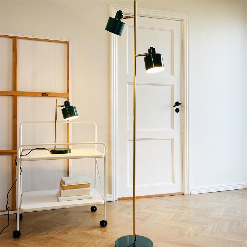 En gulvlampe med grønne lyskilder står tændt og lyser ved en hvid dør i et lyst rum; foran et hvidt rullebord med en bordlampe og nogle bøger på trægulv.