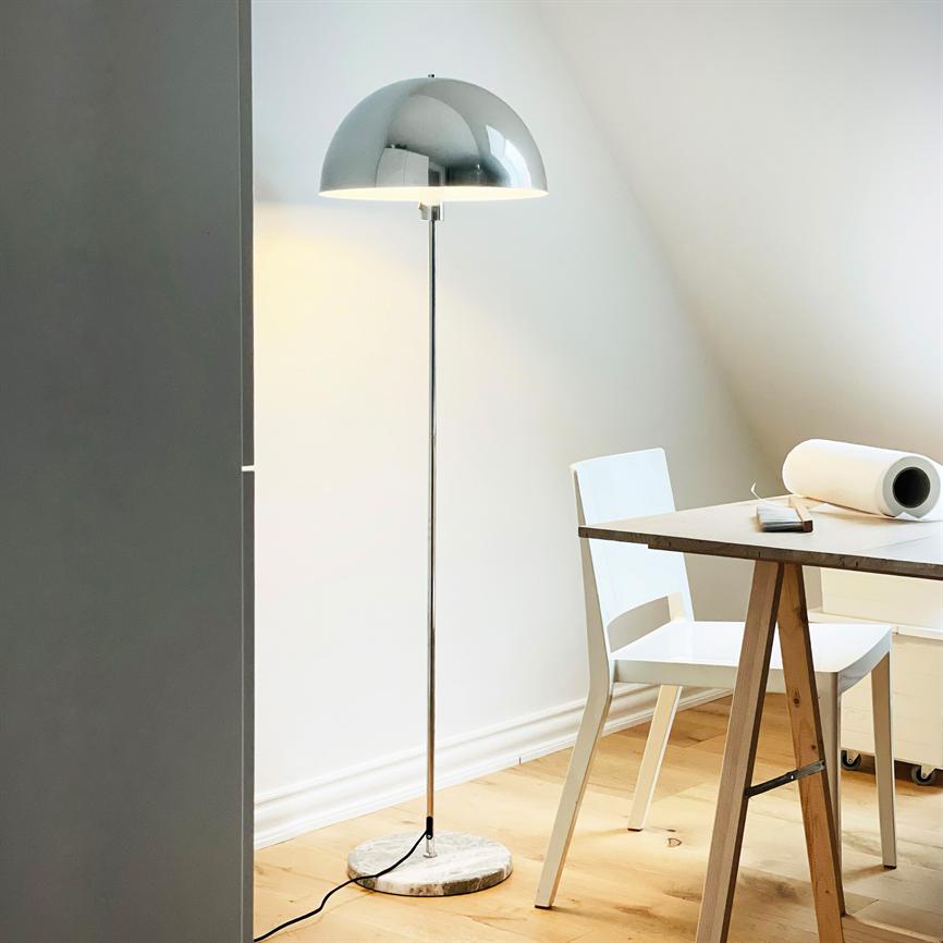Gulvlampe med blank kromkuppel står tændt på marmorfod og oplyser et hjørne med hvide vægge; ved siden står en hvid stol og et træbord med rullepapir og telefon.