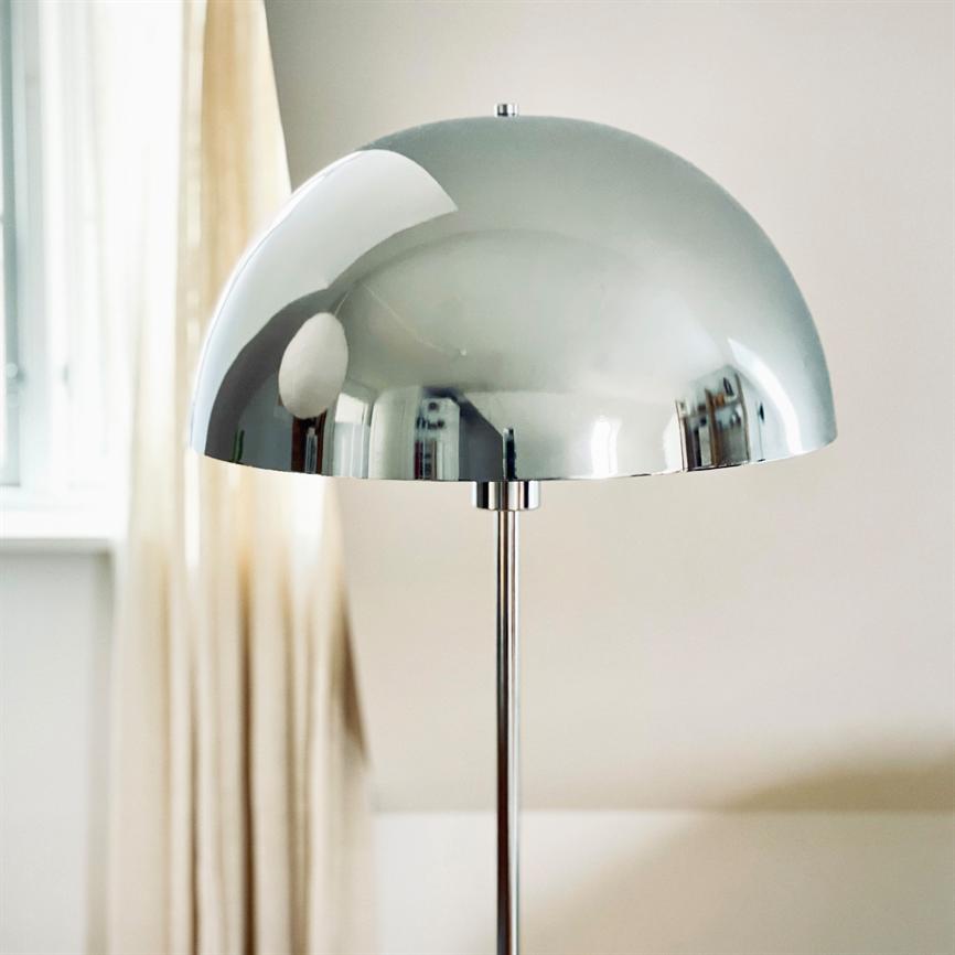 En krom halvklode-lampe reflekterer et lyst rum; den står på en slank stang ved et vindue med lyse gardiner i et neutralt, roligt interiør.