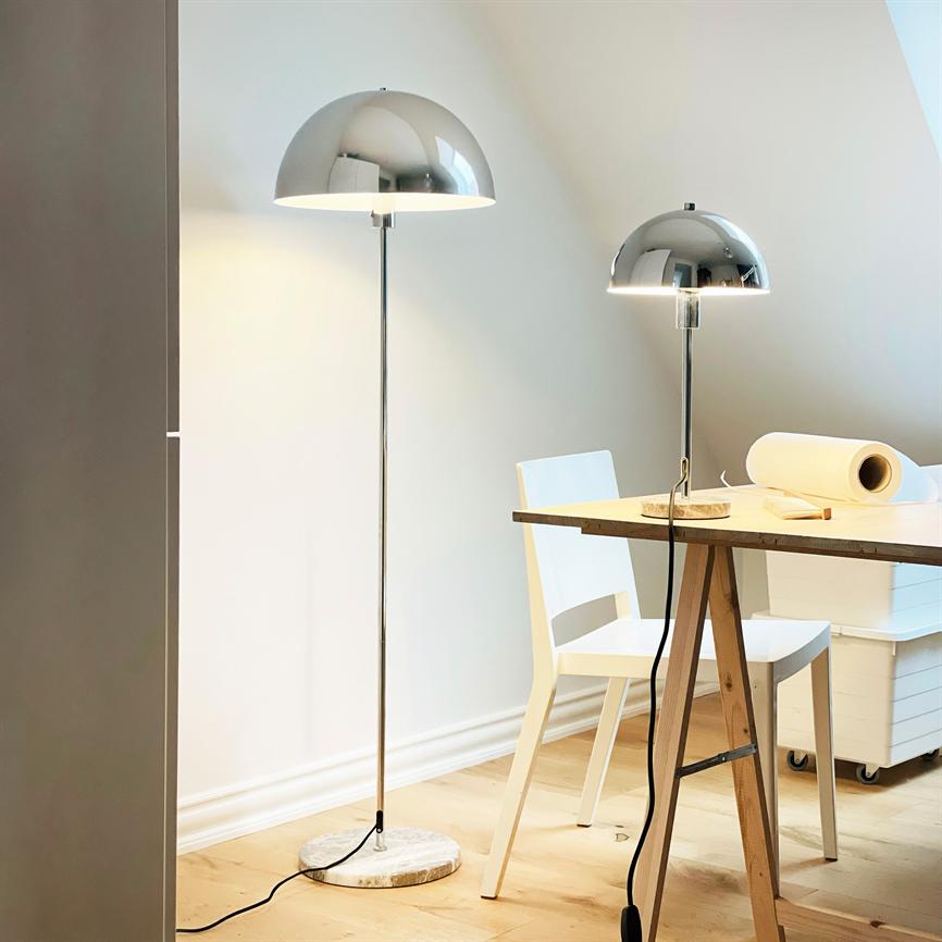To kromkuplede lamper — en stander- og en bordlampe — lyser over et træarbejdsbord med rullepapir og hvid stol i et lyst, minimalistisk rum med skråvæg og trægulv.