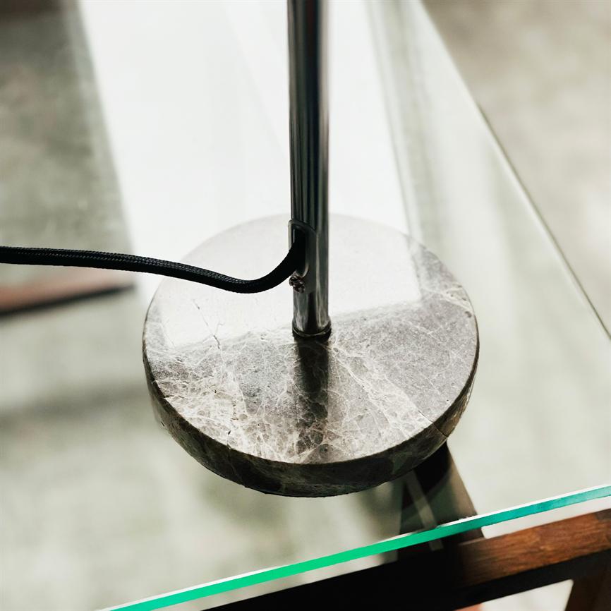 Rund marmorfod til lampe hviler på et glasbord; en kromstang stiger op fra midten, en sort tekstilledning føres ud fra stangen; baggrunden er sløret gulv.