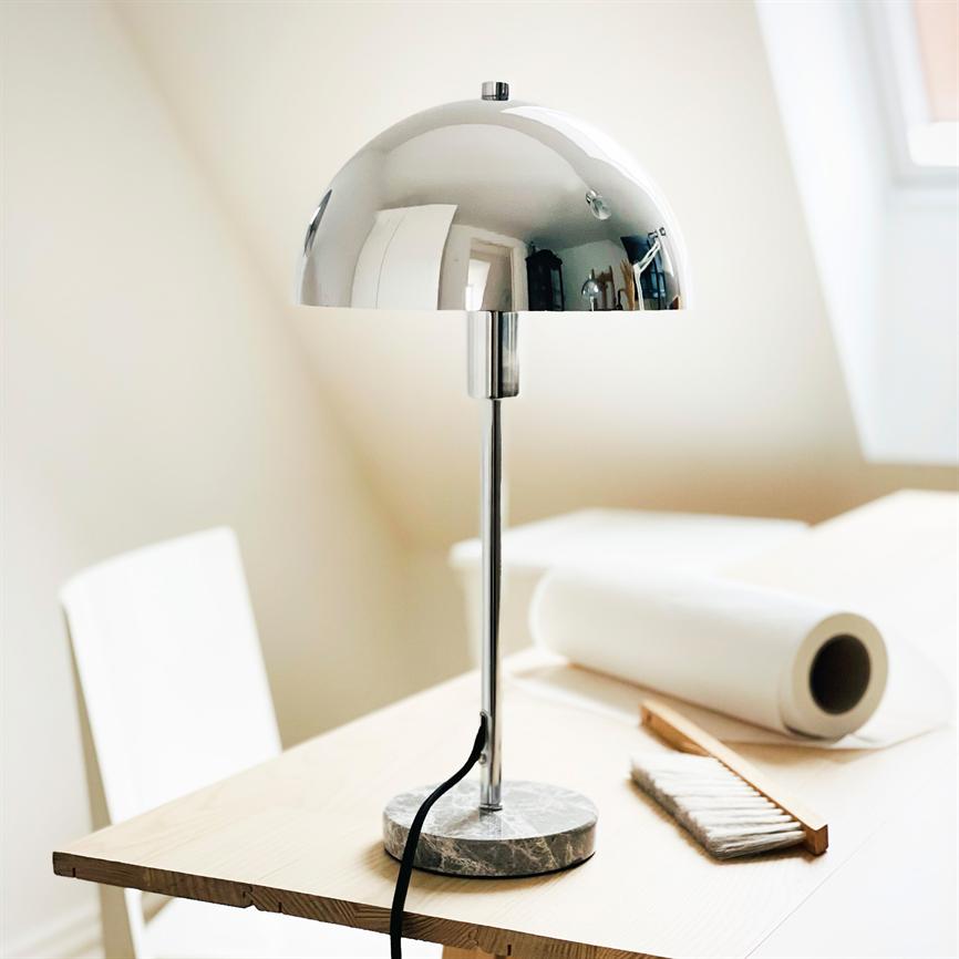En krom‑skærmet bordlampe med marmorfod står på et lyst træbord og reflekterer rummet; omgivelserne er et lyst loftsrum med en papirrulle og en lille børste ved siden af.