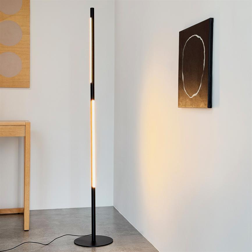 En slank sort gulvlampe udsender varmt vertikalt lys i et hjørne af et minimalistisk rum med hvid væg, betongulv, et træbord og et sort maleri med hvid oval.