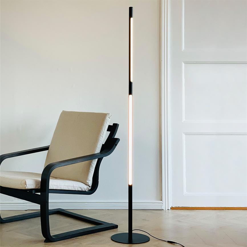 Slank sort gulvlampe udsender blødt, varmt lys ved siden af en beige polstret lænestol i et lyst rum med hvid væg og trægulv foran en hvid dør.