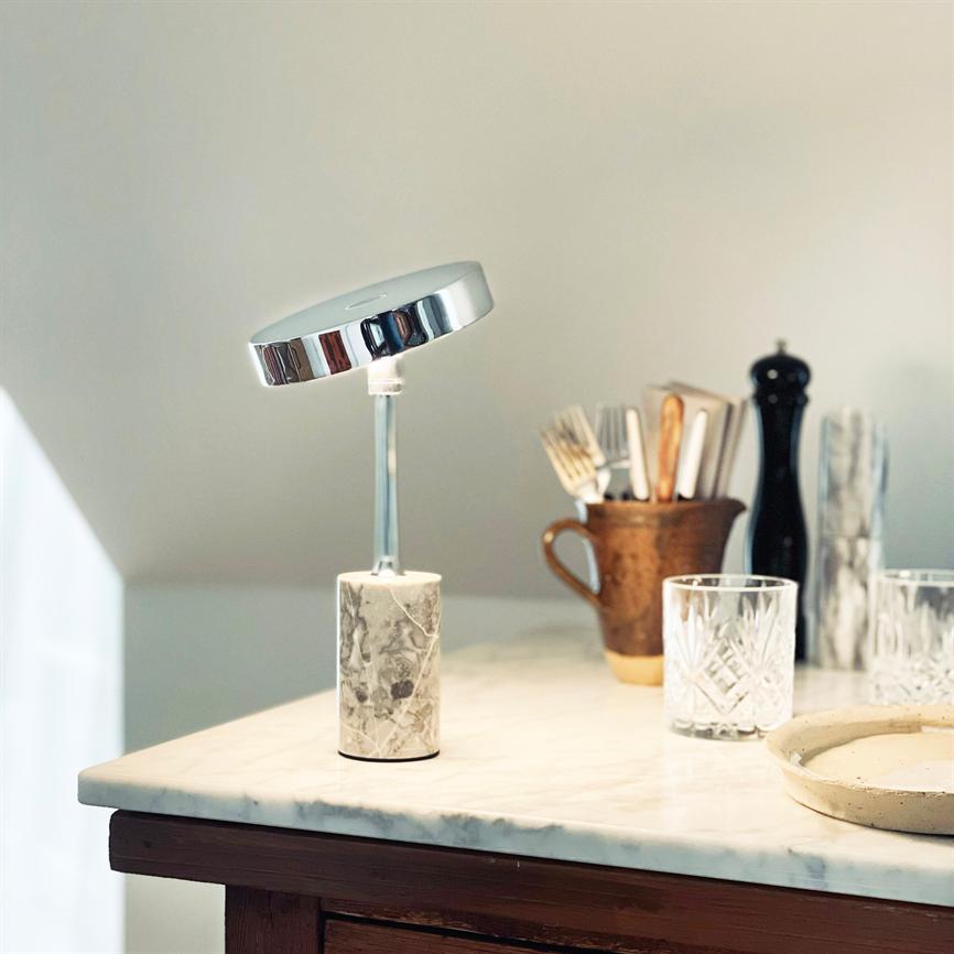 En moderne bordlampe med rund kromskærm og marmorfod står på en marmorbordplade; i baggrunden krus med bestik, glas, peberkværn og et lille fad.