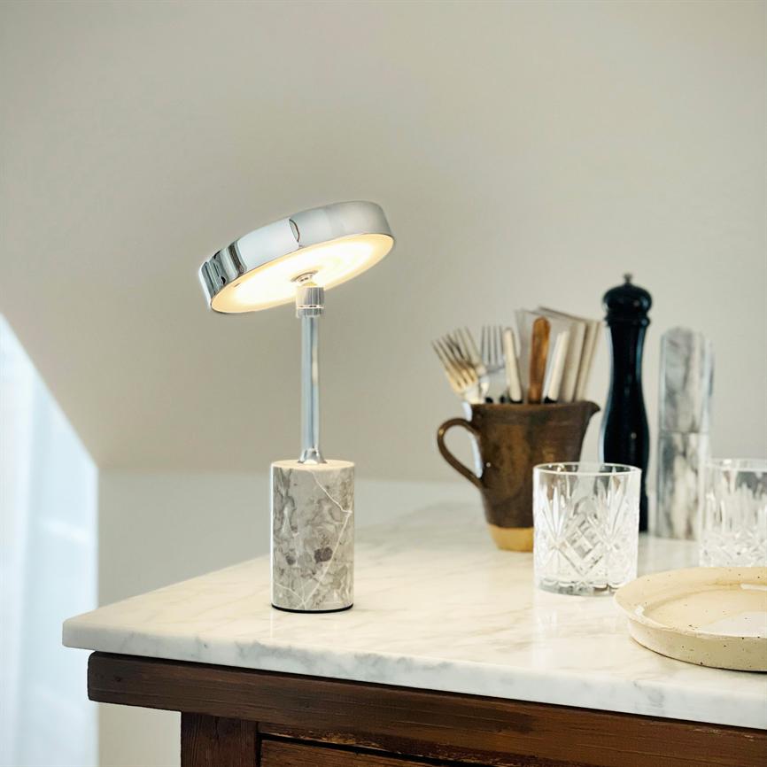 Bordlampe med rund kromskærm og marmorfod lyser blødt; står på hvid marmorbordplade i et køkken/servingsområde med glas, bestik i krukke og peberkværn i baggrunden.