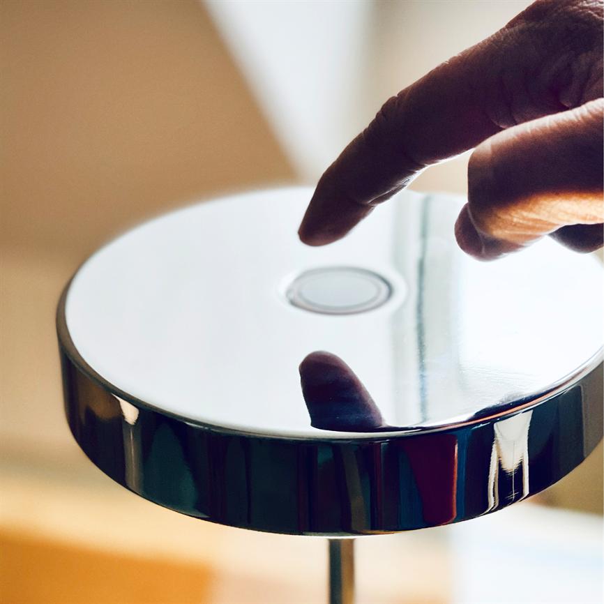 En blank, cirkulær lampeoverflade; en fingerspids nærmer sig og reflekteres i den; lampen står på en kromstander i et varmt, oplyst indendørs rum.