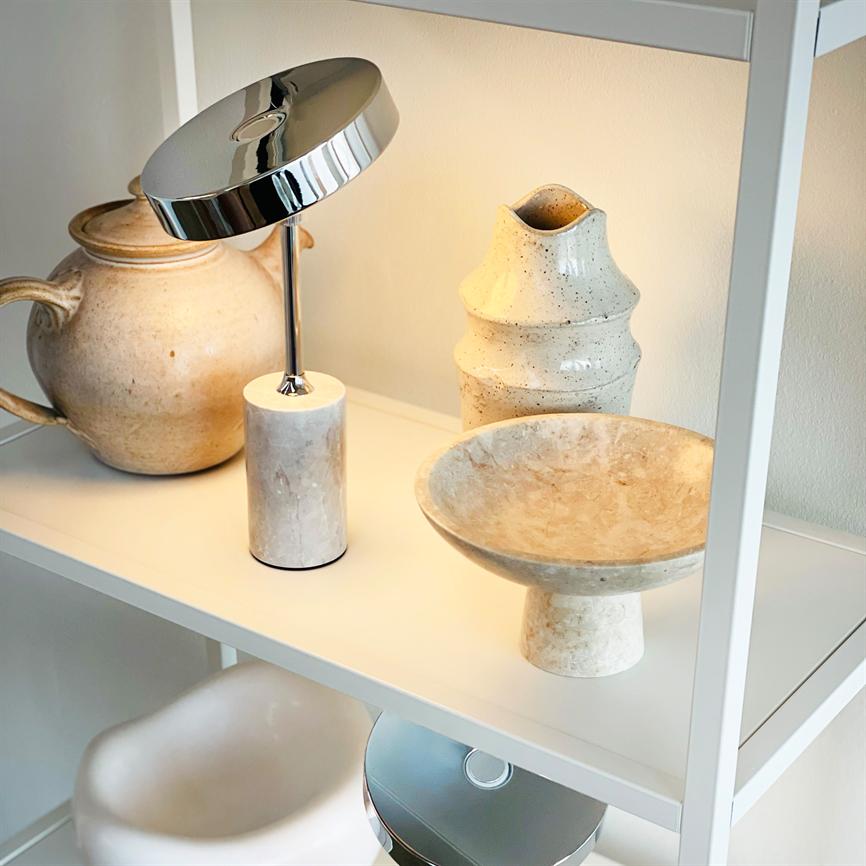 Kromlampe står tændt og kaster varmt lys på en hvid hylde; omkring den står støvede beige keramikkrukker og en skål på fod i en neutral, minimalistisk indretning.
