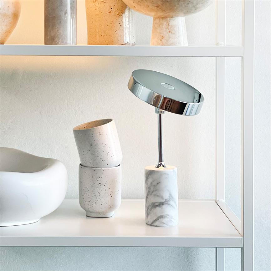 Bordlampe med rund kromskærm vipper på en slank stang over en marmor-cylindrisk base, placeret på en hvid hylde ved siden af to stabelbare keramikkrus og en skål.