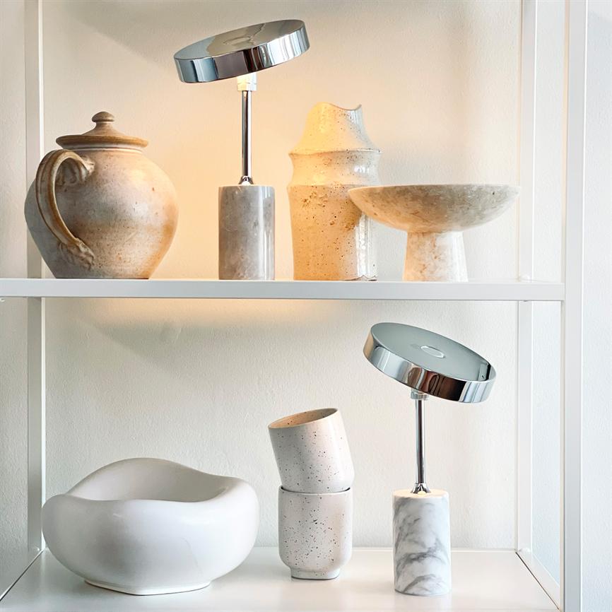 Kromhovedet bordlampe med marmorfod kaster blødt, varmt lys mellem rustikke beige keramikkrukker, vaser og skåle på hvide hylder i et lyst rum.