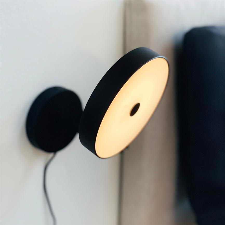 Væglampe udsender varmt, diffust lys; monteret på hvid væg med synlig sort ledning; placeret ved en beige sengegavl og et mørkt stofmøbel.