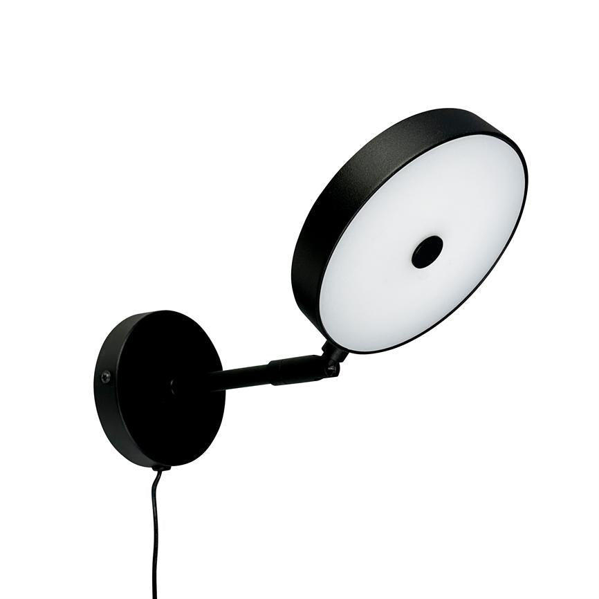 Rund væglampe med sort metalramme og hvid diffusor; vippet udad på en kort arm fra en rund vægplade, med synlig sort ledning, mod neutral hvid baggrund.