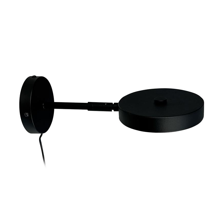 Sort væglampe med rund vægbasis; en lige arm fører til et fladt, cirkulært lampehoved, ledning hænger ned, mod neutral hvid baggrund.