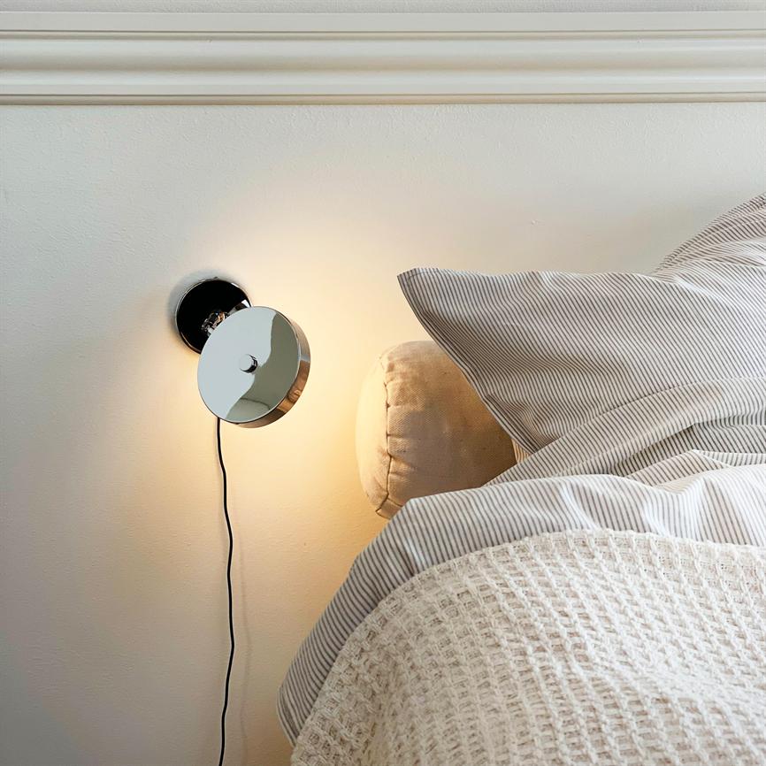 Væglampe lyser svagt over en seng; krom rund lampe med synlig ledning, beige hovedpude og stribet lagen samt en lys, vævet dyne mod en neutral væg.